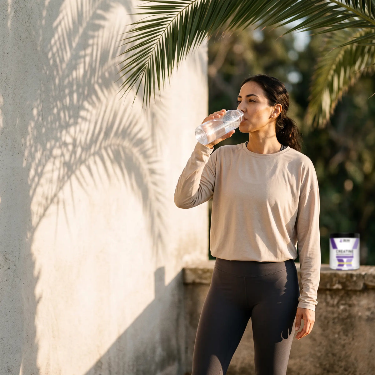 A woman drinking water on a shaded terrace after training, with palm shadows on the wall behind her