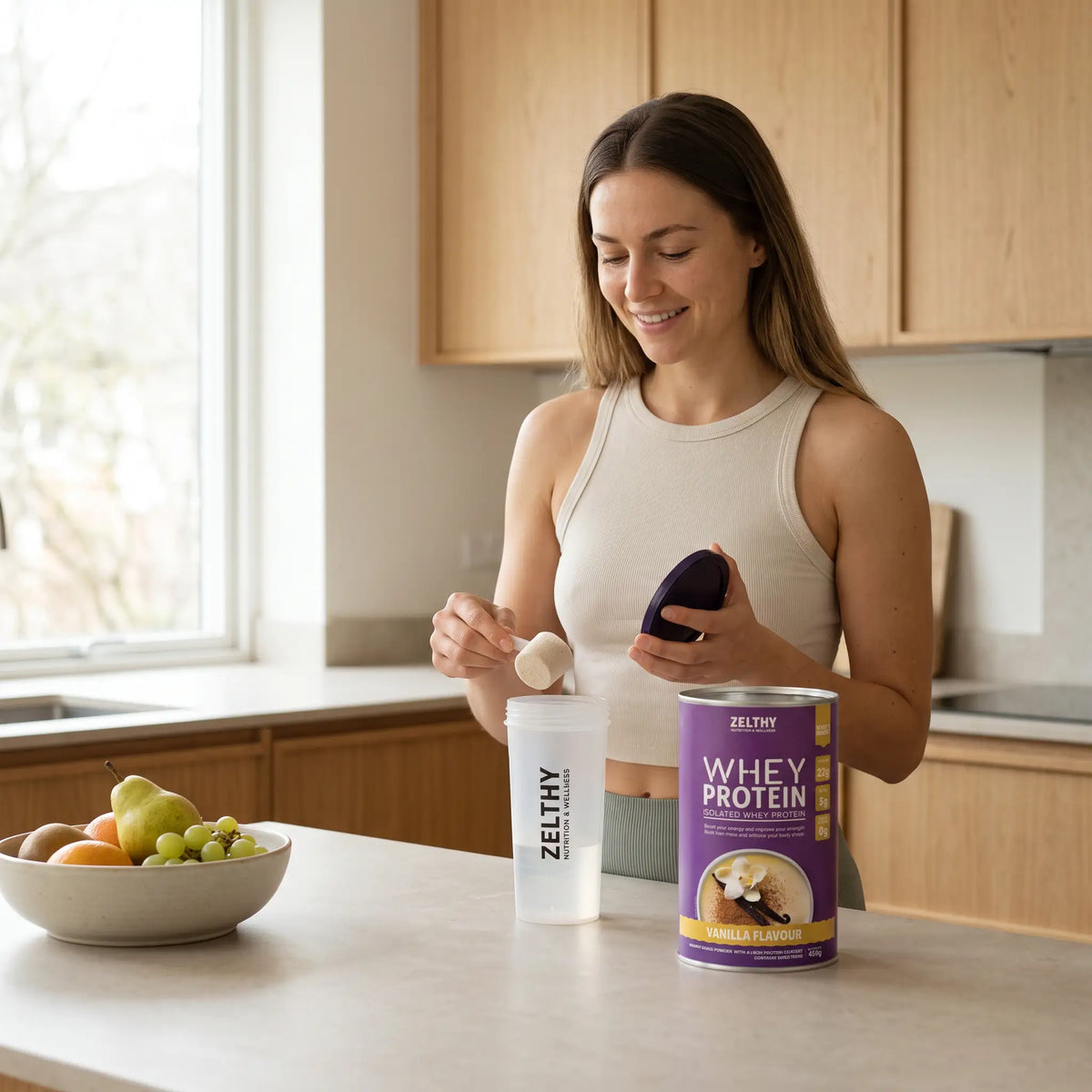 A woman in a light kitchen scooping vanilla Zelthy Whey Protein from the 450 g tub into a shaker