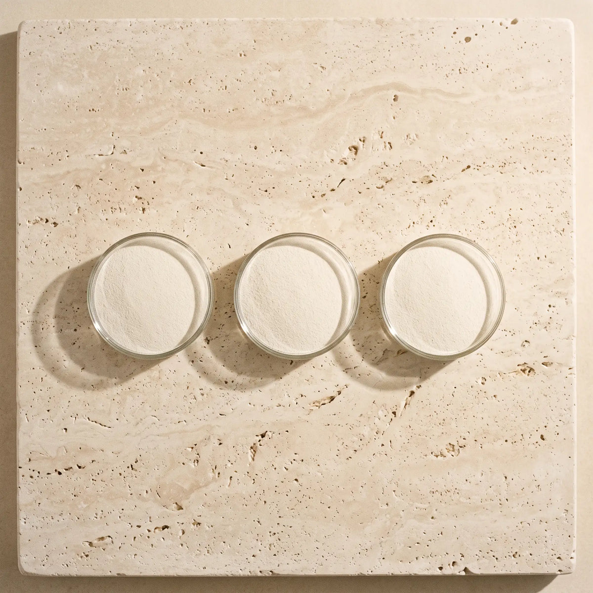 Three glass dishes on a travertine slab, each holding an identical level measure of unflavoured supplement powder