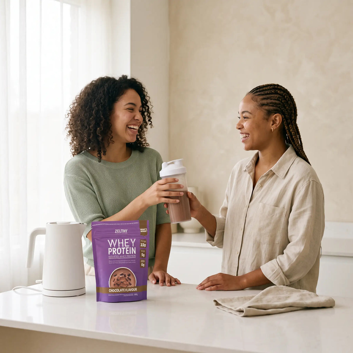 Two friends in a bright kitchen sharing a chocolate protein shake next to a pack of Zelthy Whey Protein